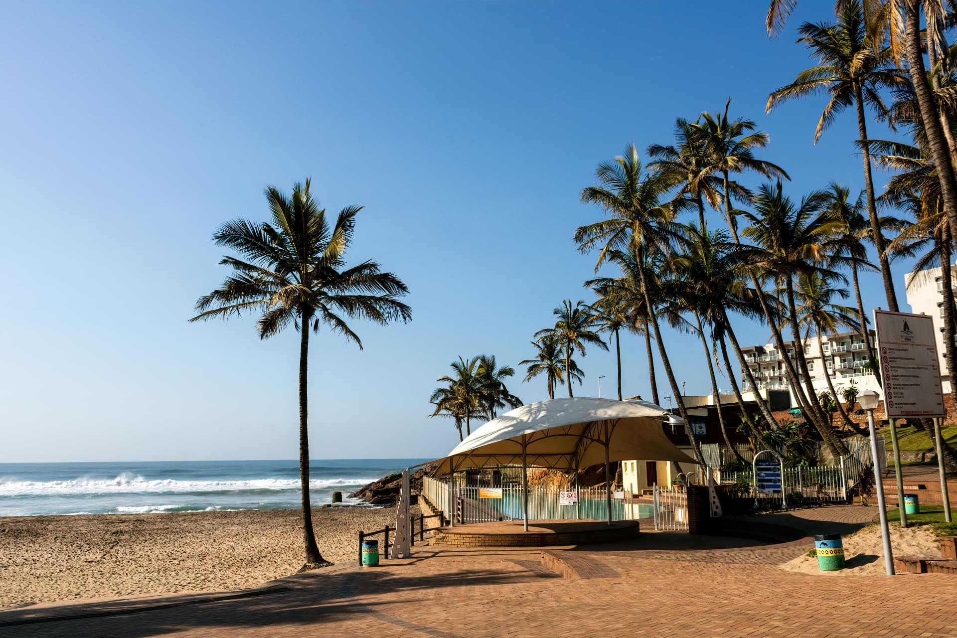 The palm-lined Ballito promenade at Willard Beach, its paved walkway leading past the tented pavilion above the tidal pool